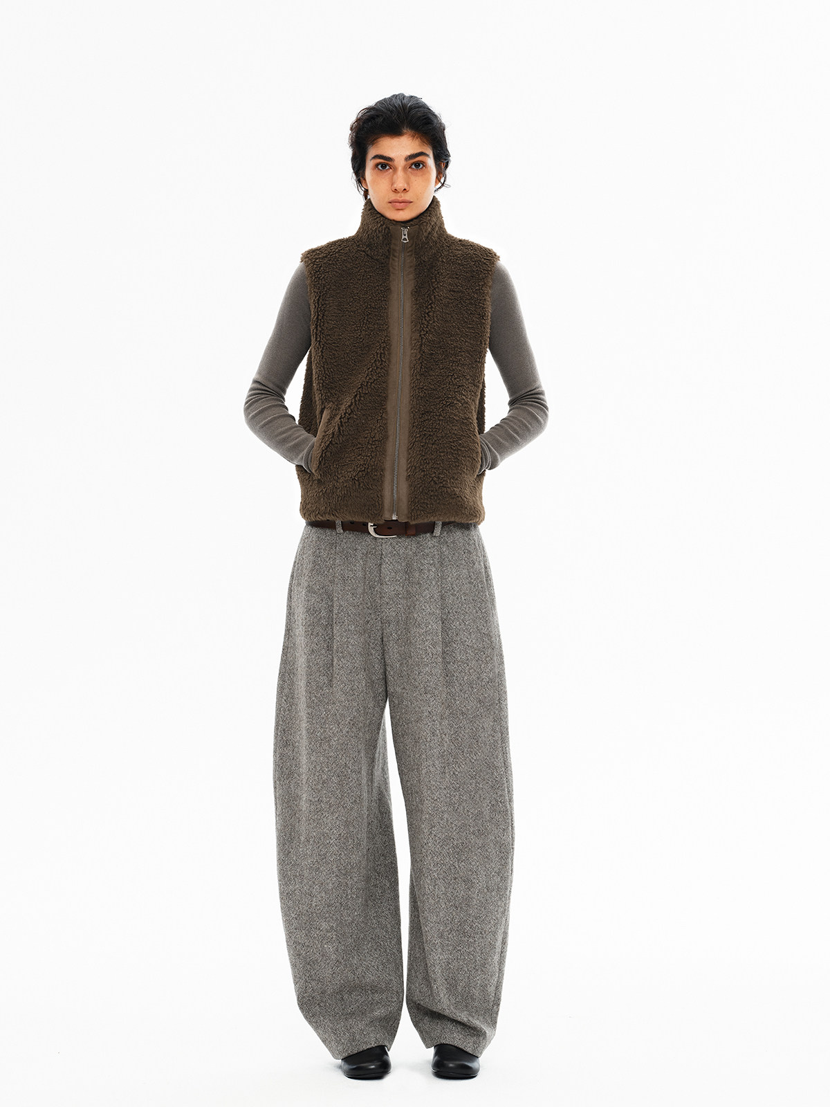 一个人站在纯白色背景前，面向前方，身穿 EastLyst 高领羊羔绒背心，内搭灰色长袖衬衫，下身穿着宽松的灰色长裤和黑色鞋子，双手插在口袋里。
