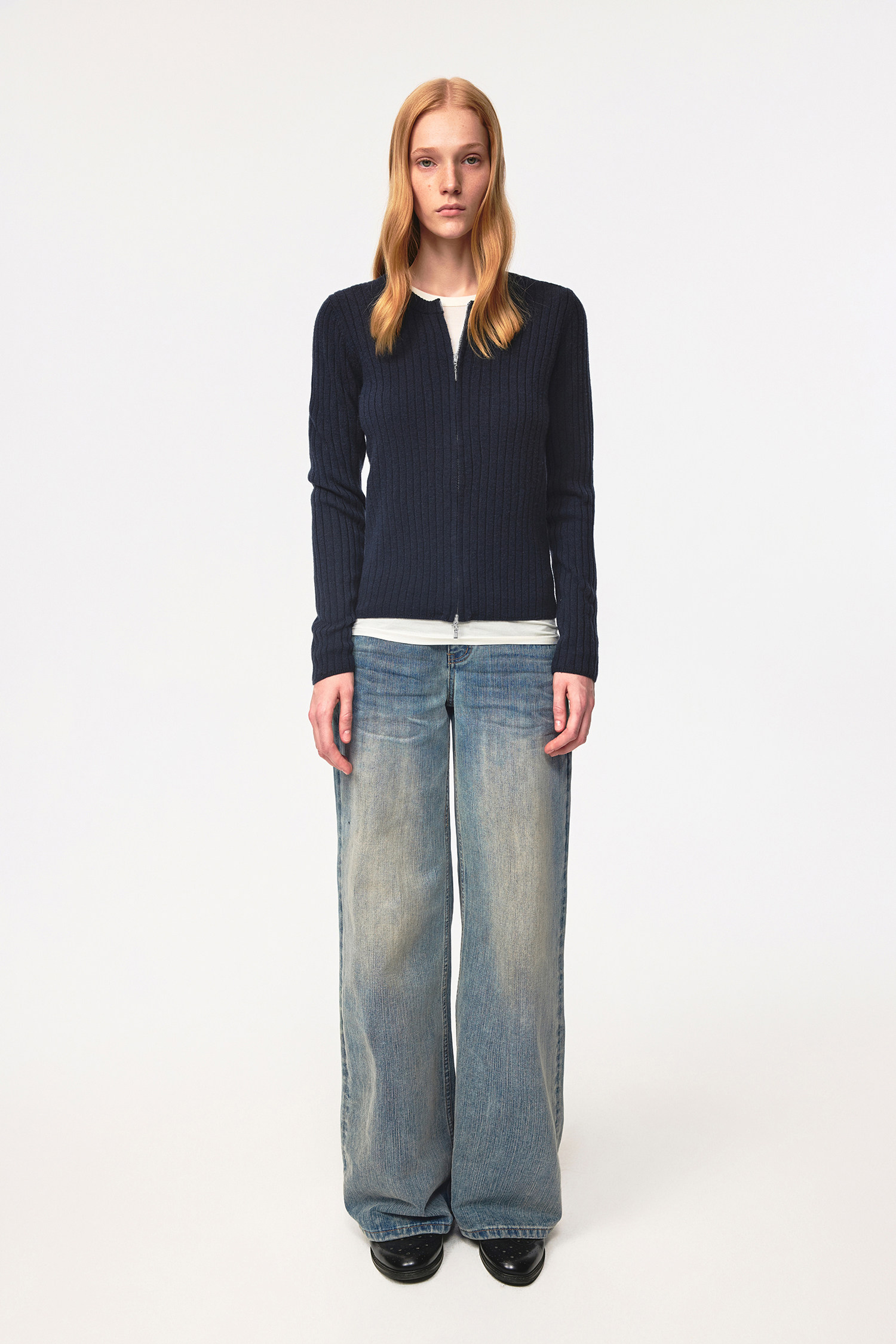 A person with long blonde hair faces forward, wearing an EastLyst Merino Wool Cardigan in navy over a white shirt, paired with wide-leg faded blue jeans and black shoes, standing against a plain white background.