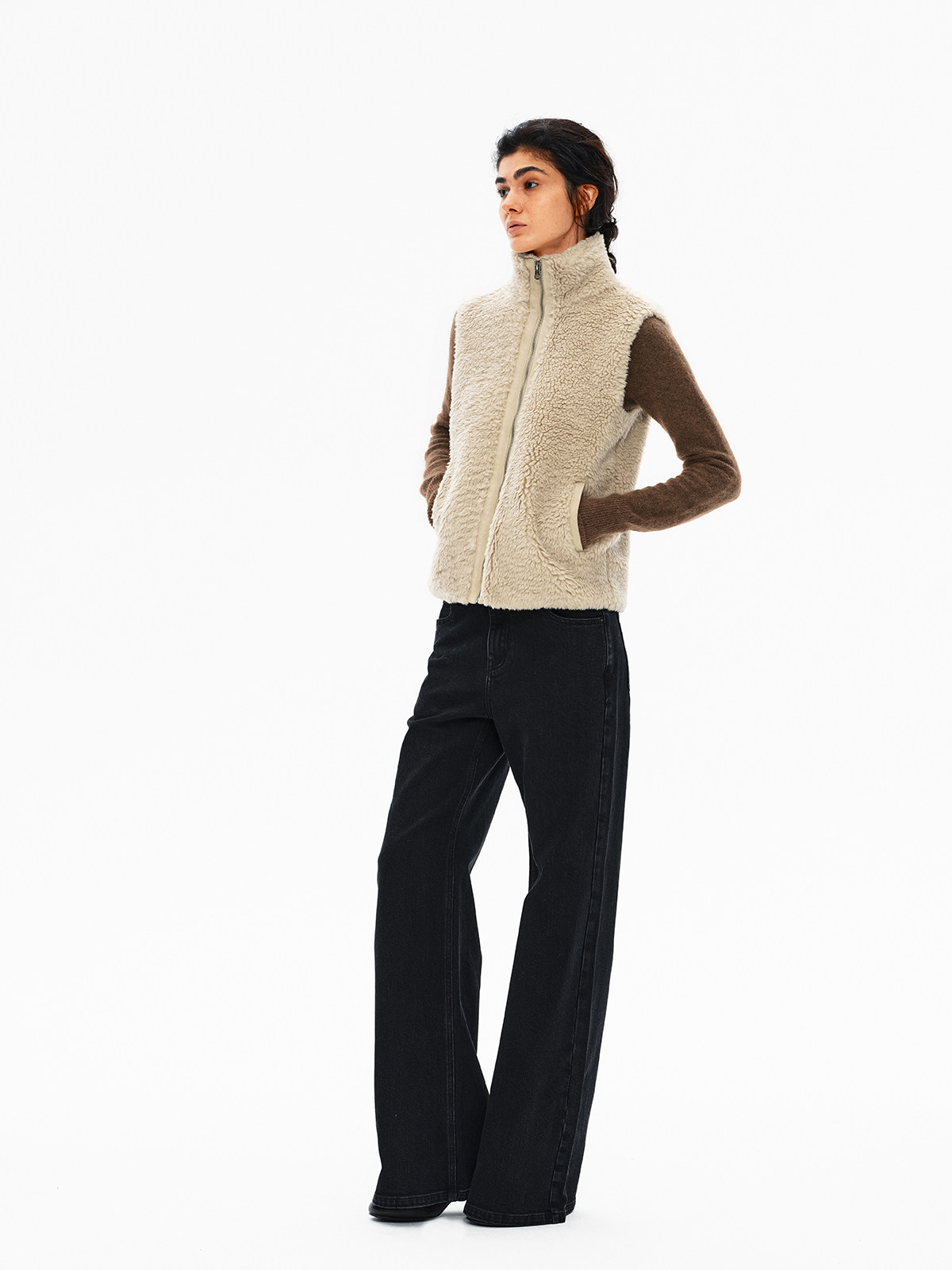 在白色背景下，一个人穿着 EastLyst 高领羊羔绒背心，内搭棕色长袖上衣和黑色阔腿牛仔裤，双手插在背心口袋里，目光略微侧向一边。