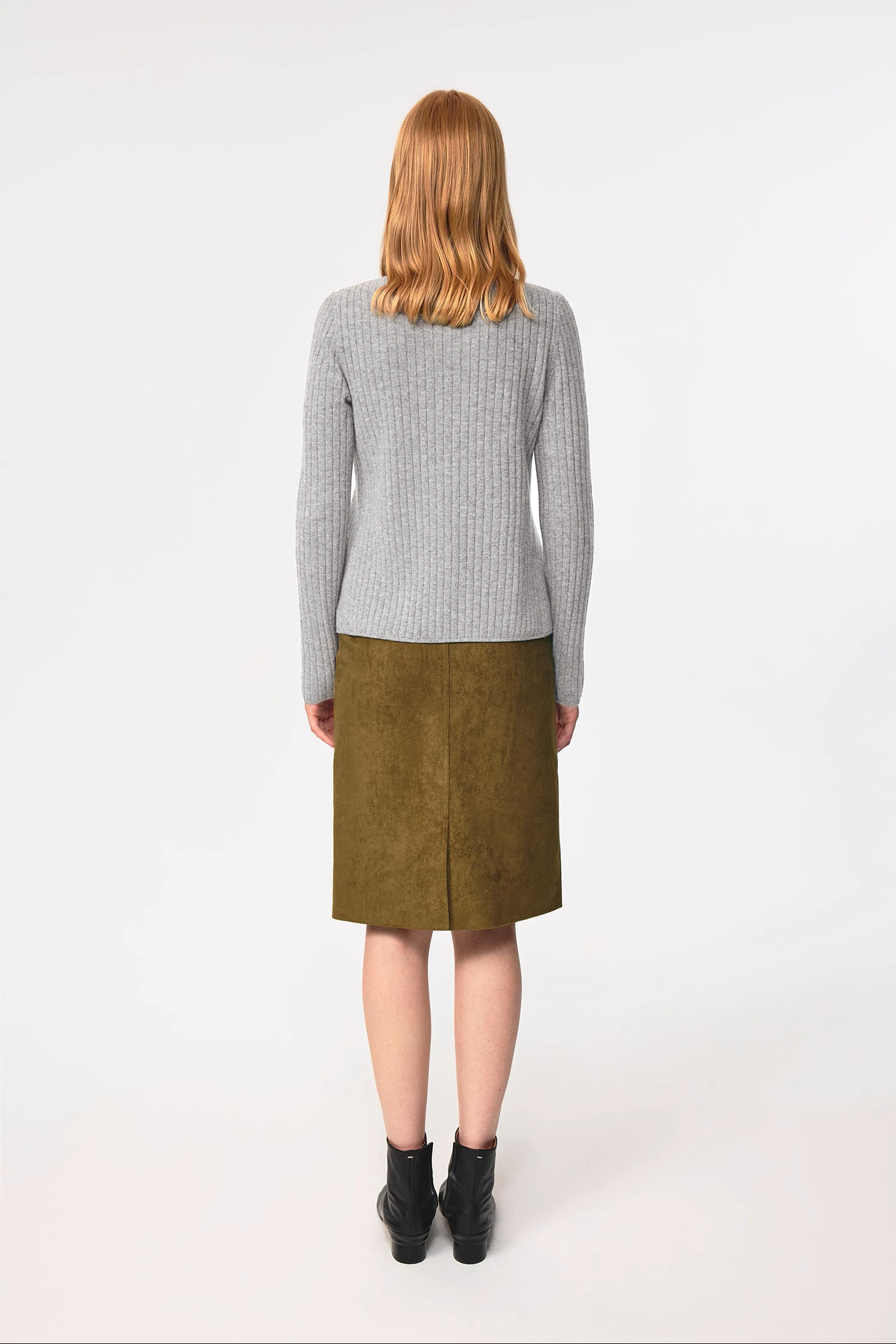 A woman with shoulder-length auburn hair stands facing away, wearing the EastLyst Merino Wool Cardigan in gray with an olive green skirt and black ankle boots against a plain white background.