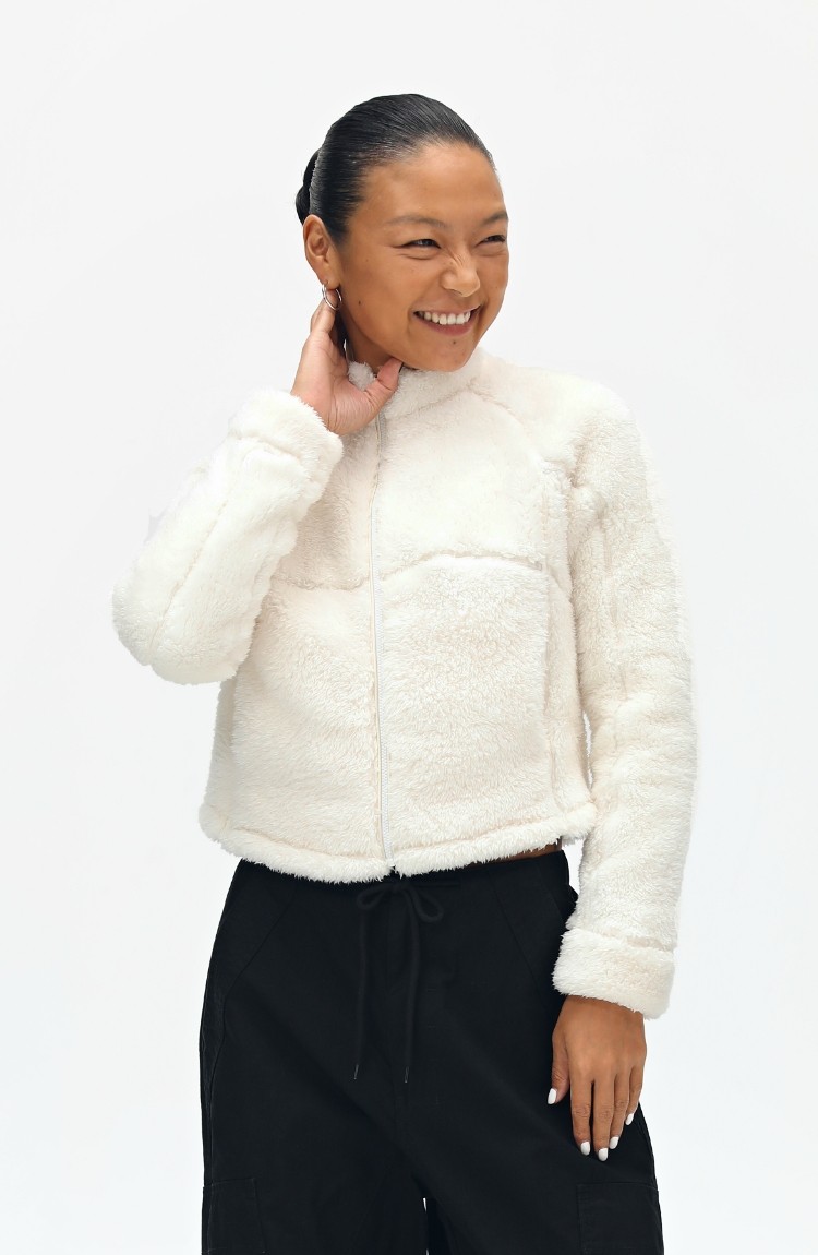 A person wearing the EastLyst Pink Short Faux Shearling Jacket and black pants smiles while touching their neck, standing against a plain white background.