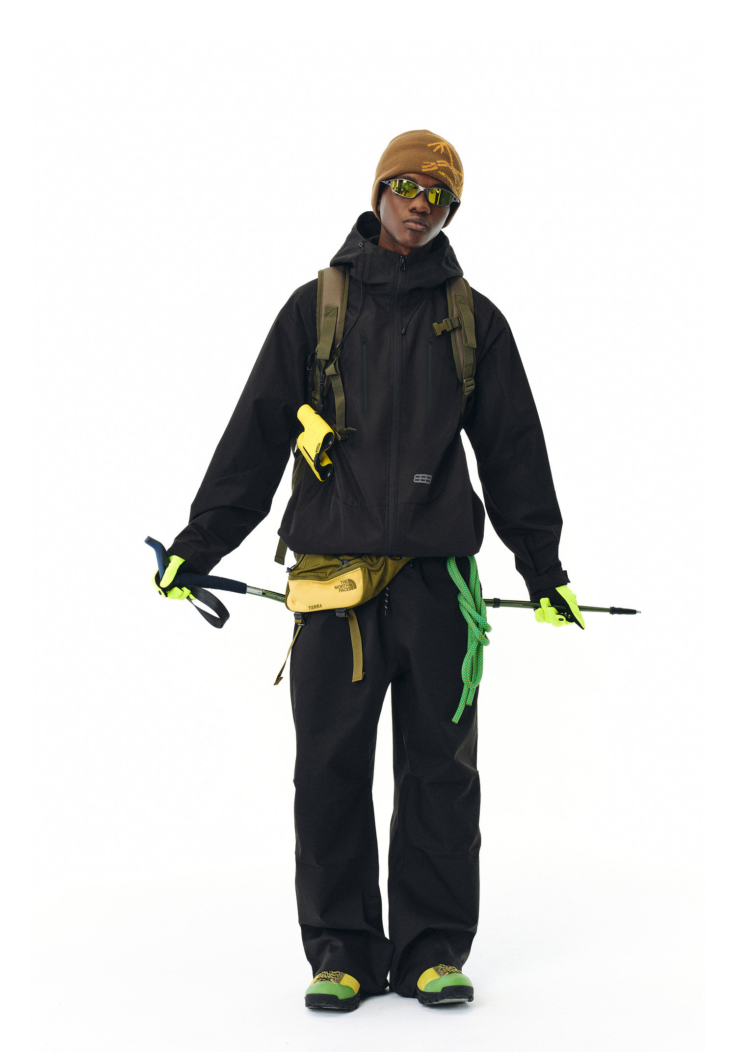 Outfitted in the EastLyst Best All-Rounder Jacket, this hiker sports outdoor pants, neon-accented boots, a brown beanie, reflective sunglasses, a backpack, and grips trekking poles.