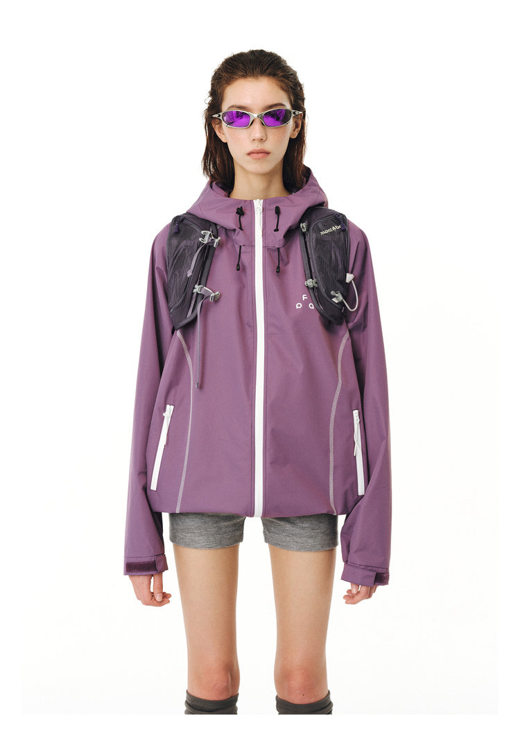 A person faces forward against a plain white background, wearing an EastLyst protective outdoor jacket in purple with a hood, gray shorts, purple-tinted sunglasses, and a small black backpack on their chest.
