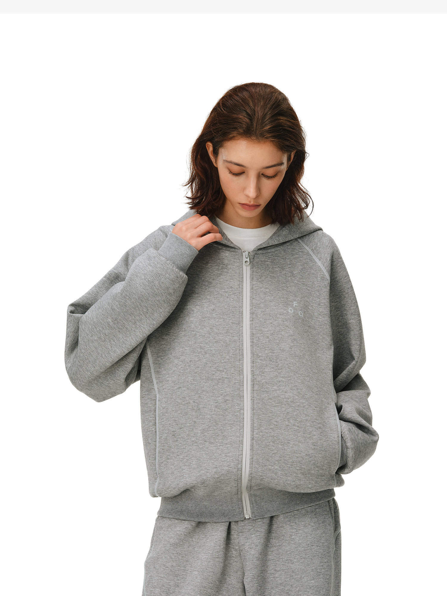 A person with shoulder-length brown hair wears EastLyst’s Constant Warmth Zip Hoodie and matching sweatpants, crafted from high-end fabric, standing against a white background and looking down while holding the hoodie zipper.