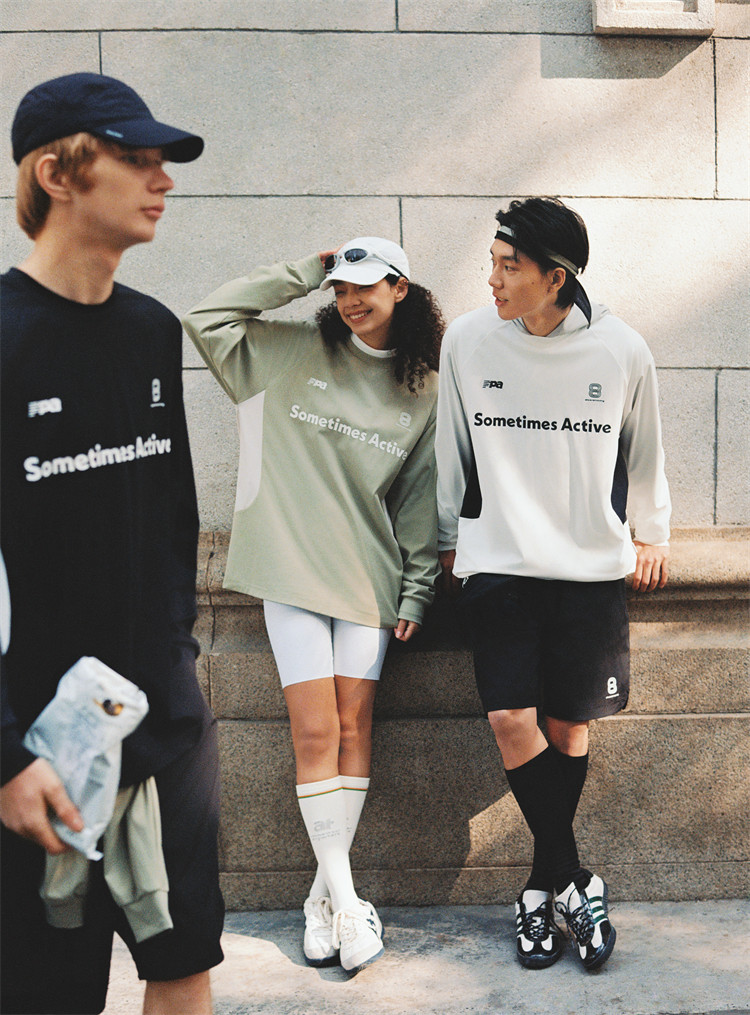 Three young adults in EastLyst Quick-Dry Color Block Long Sleeve Tees chat outdoors by a stone wall. Two lean and smile at each other, while another walks by with a bag; all shirts feature "Sometimes Active" printed on the front.