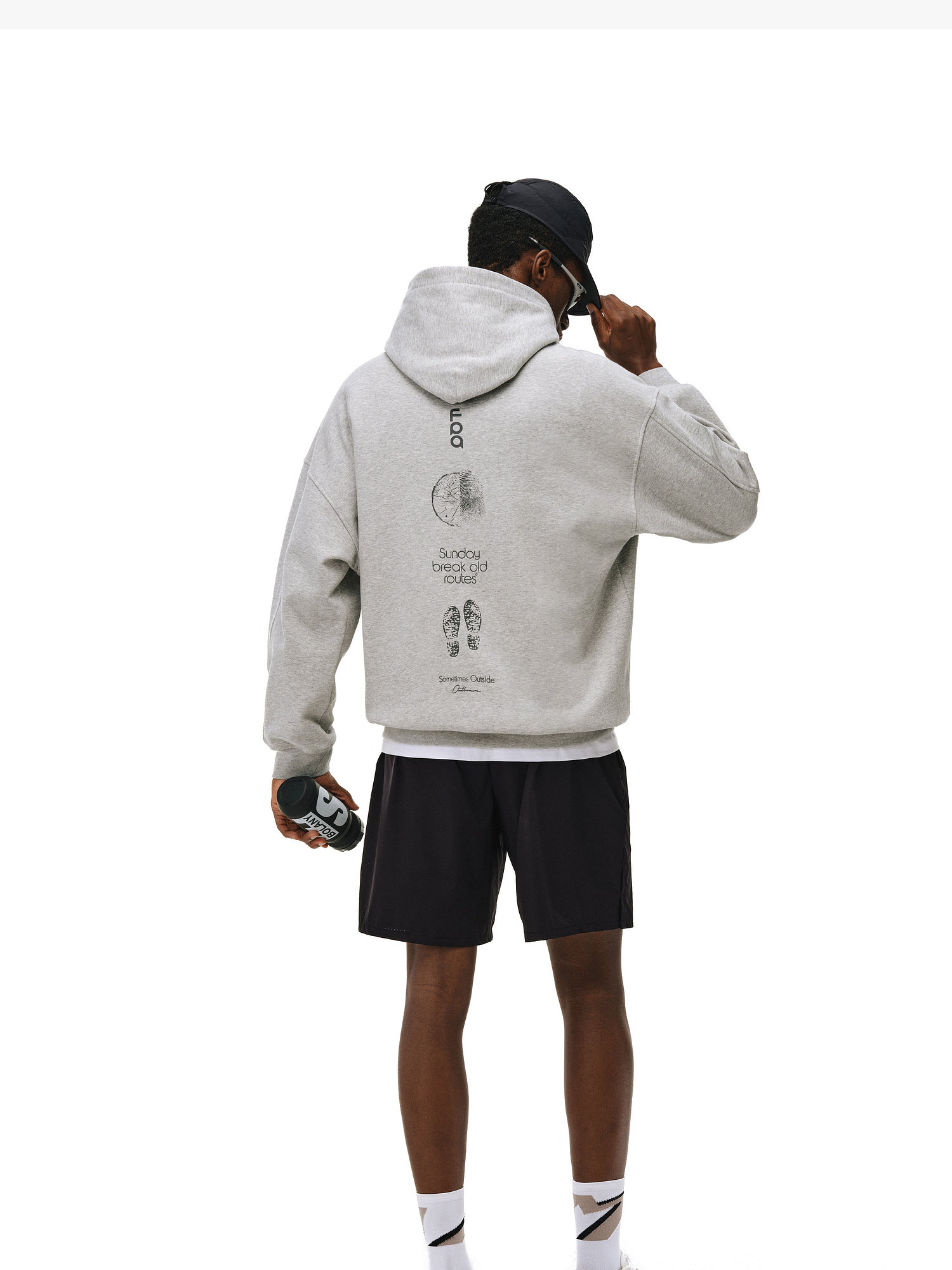 A person faces away, wearing the EastLyst Outdoor Print Hoodie with black graphics on the back, paired with black shorts, white socks, sneakers, and a black cap while holding a water bottle. The background is plain white.