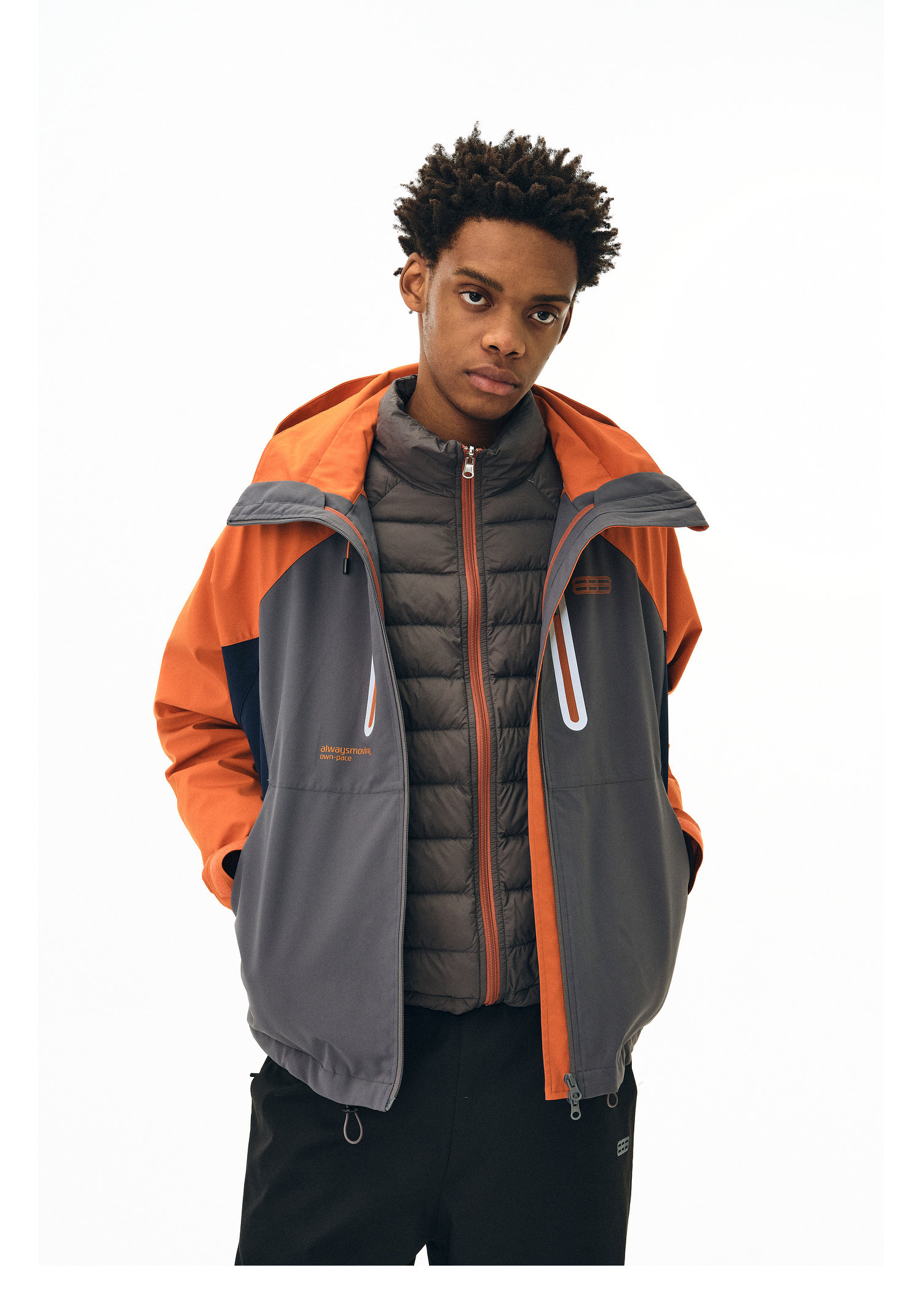 A person stands against a plain background wearing the EastLyst Versatile Explorer 3-in-1 Jacket in gray and orange over a black zip-up vest, hands in their pockets with a neutral expression.