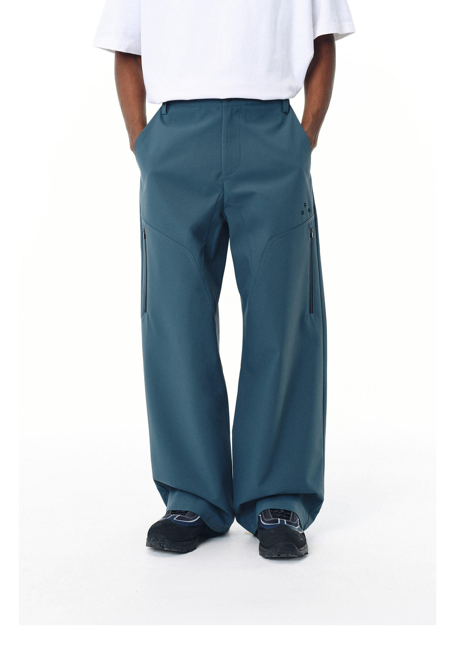 A person wears EastLyst Waterproof Windproof Softshell Pants—loose-fitting, teal, with side zippers—paired with a white shirt. Only the lower body is shown against a white background, hands in pockets.
