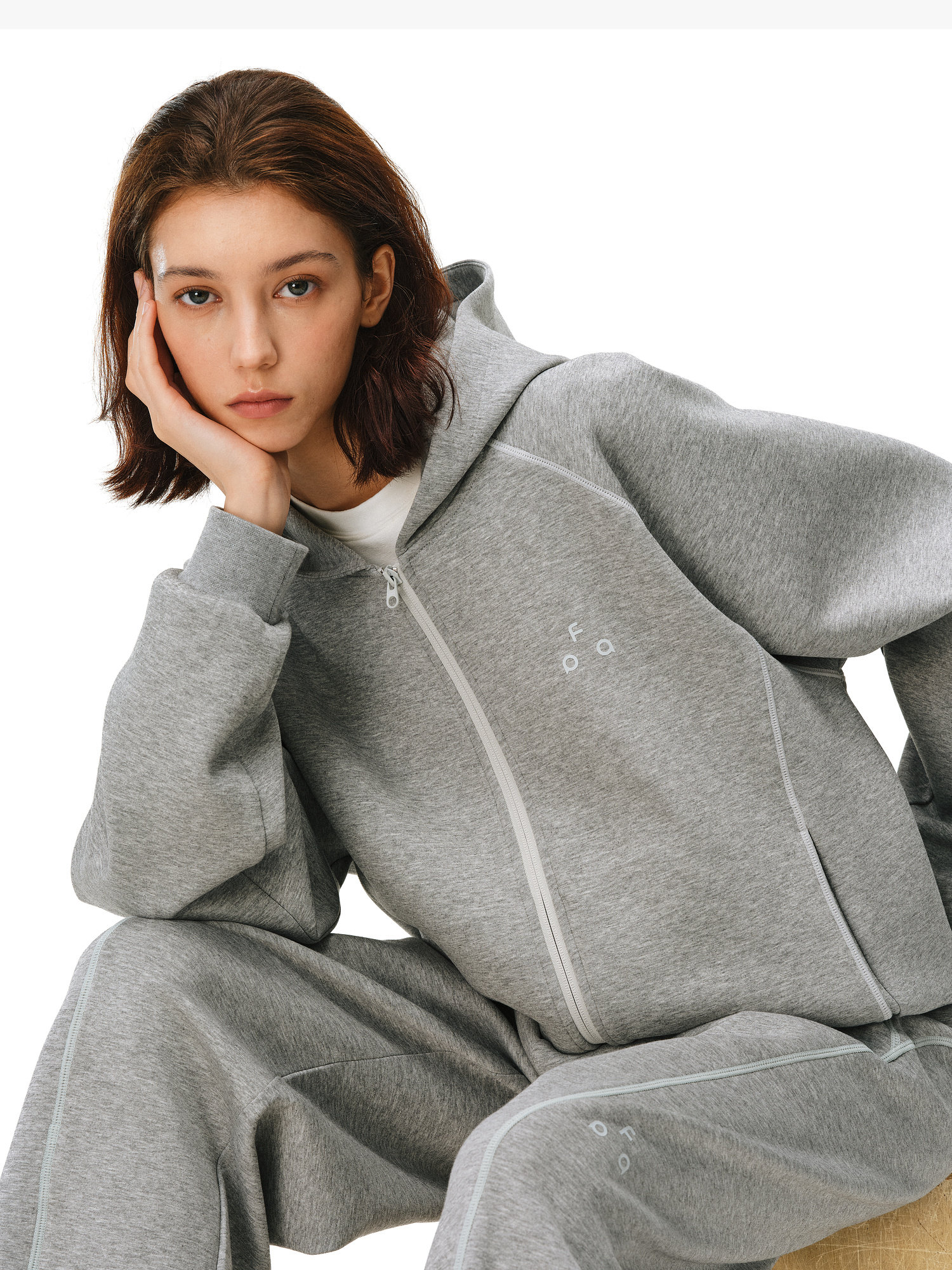 A young woman with brown hair wears the EastLyst Constant Warmth Zip Hoodie and matching sweatpants in premium fabric, sitting with her head on her hand and looking directly at the camera with a neutral expression.