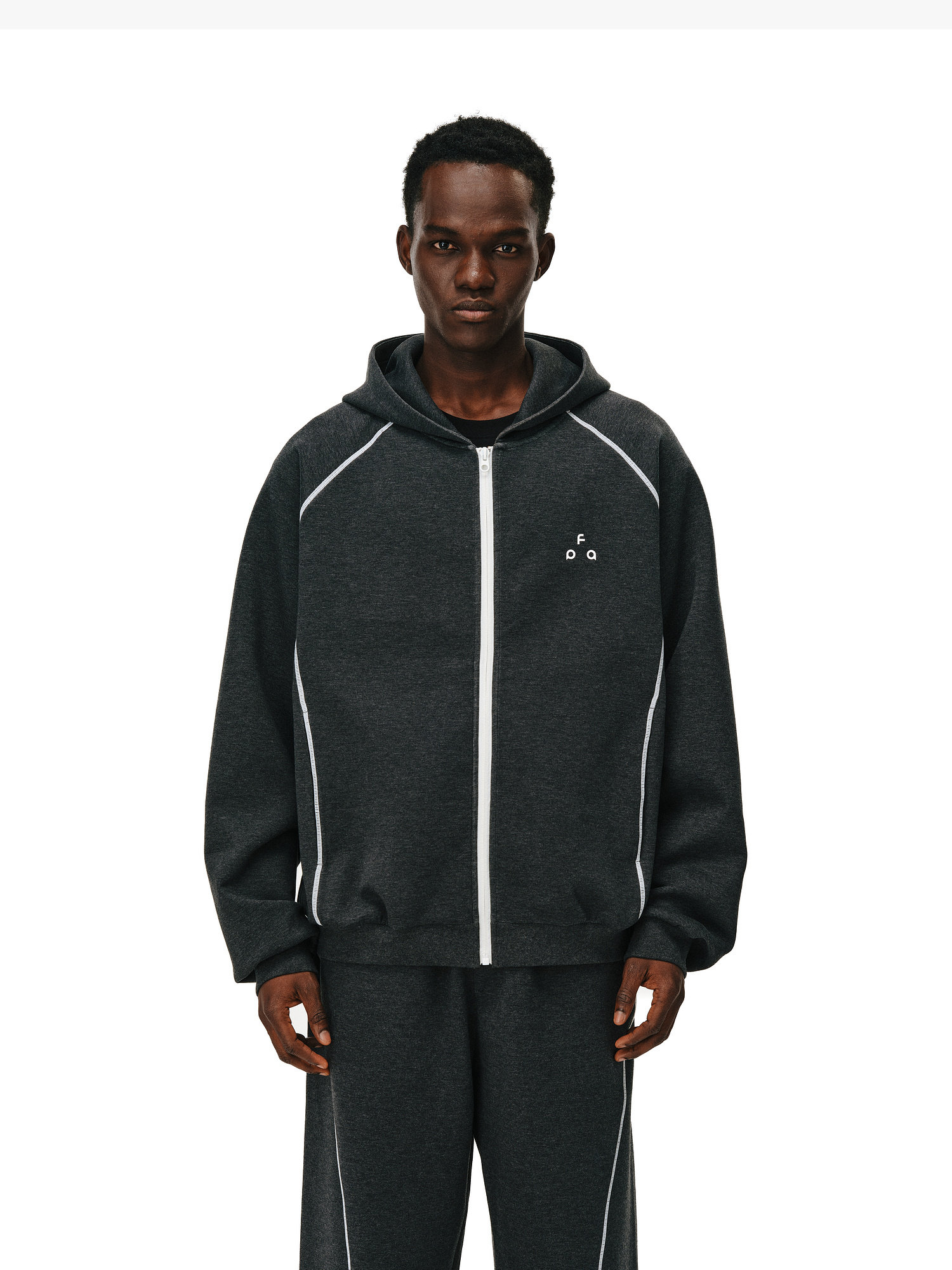 A man wears the EastLyst Constant Warmth Zip Hoodie in dark gray with FG on the chest and matching pants featuring white piping, standing facing forward against a plain white background.