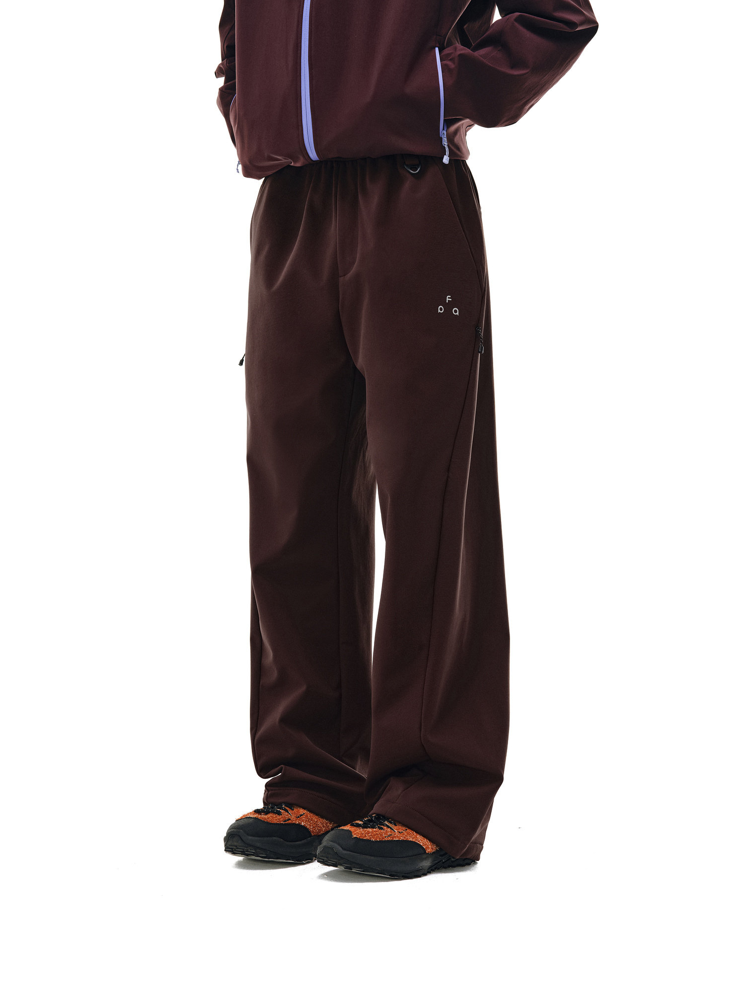 A person models EastLyst's Urban Outdoor Softshell Pants in maroon, paired with a matching jacket with purple trim and black-and-orange shoes, standing against a white background. Only the lower body is visible.