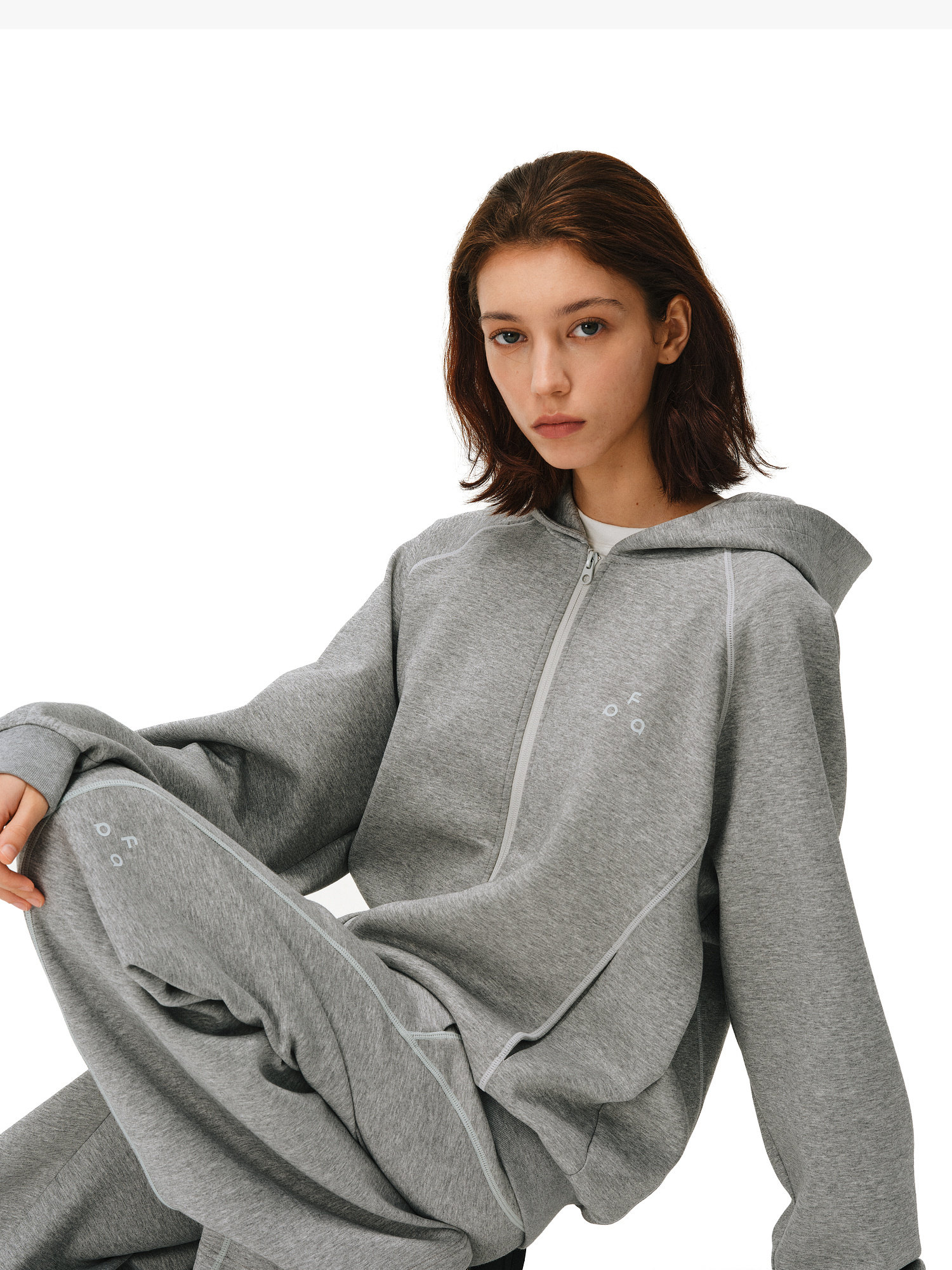 A young woman with shoulder-length brown hair wears the EastLyst Constant Warmth Zip Hoodie and matching sweatpants, crafted from premium fabric, as she sits with one knee raised against a plain white background.