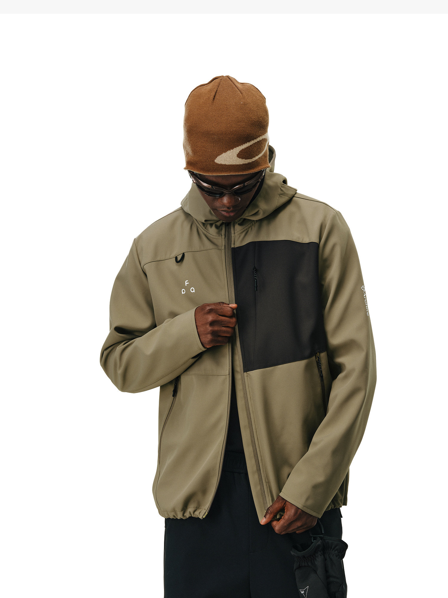 A person wearing a brown beanie, sunglasses, and the EastLyst Color-Block Water-Resistant Softshell Jacket with a black chest pocket stands against a white background, looking down while zipping up the jacket.