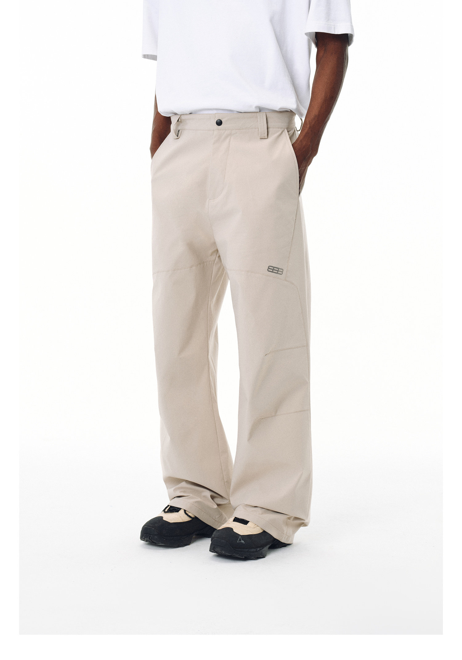 Model wearing EastLyst Outdoor water-repellent casual trousers in light beige, paired with a white t-shirt and black-and-beige shoes, standing hands in pockets against a plain white backdrop; head not visible.