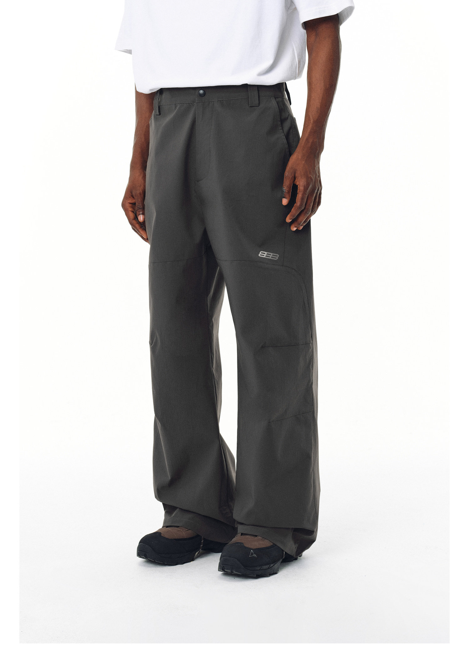 A person is shown from the shoulders down wearing EastLyst Outdoor water-repellent casual trousers in dark gray, featuring a small rectangular logo, paired with a white T-shirt and black shoes against a white background.