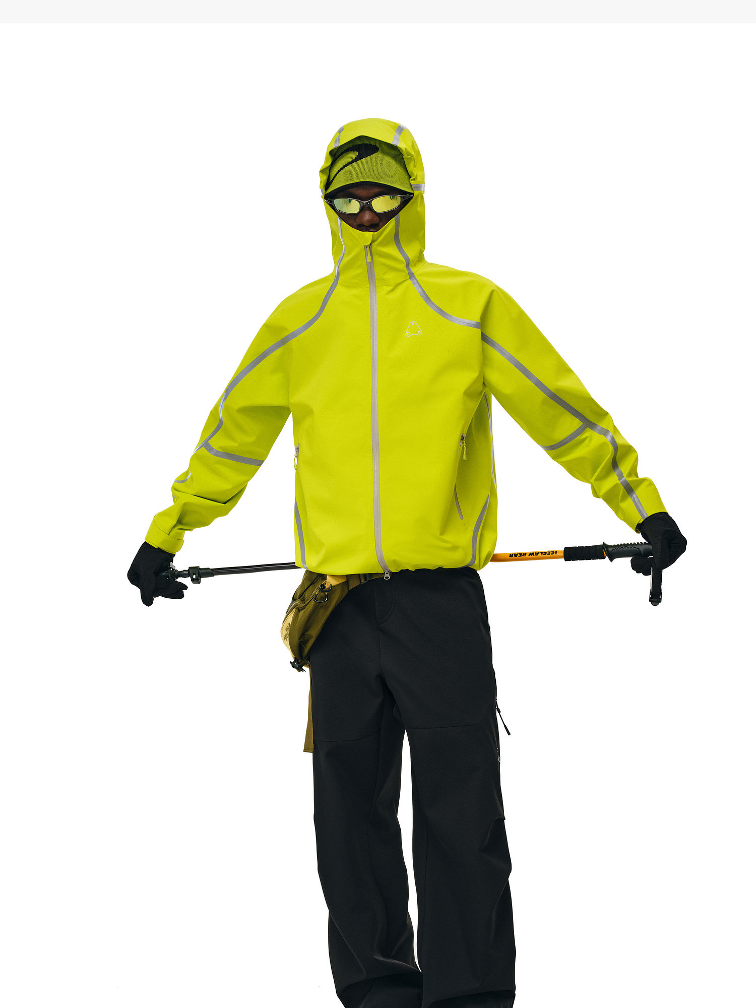 Wearing EastLyst Softshell Joggers, gloves, and sunglasses, a person in a bright yellow waterproof hooded jacket stands holding trekking poles against a white background.