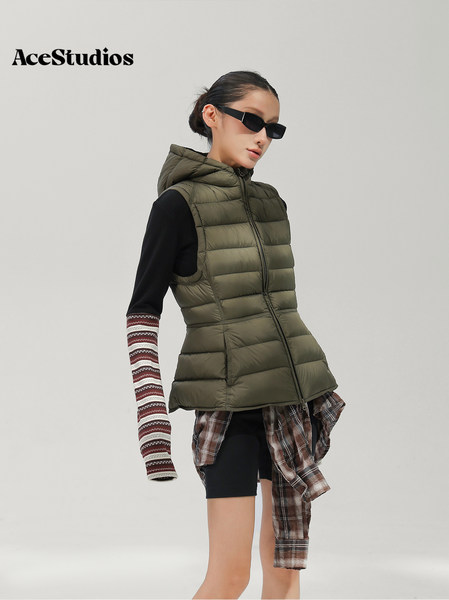 Model wearing an olive quilted hooded vest layered over a black top with plaid accents.