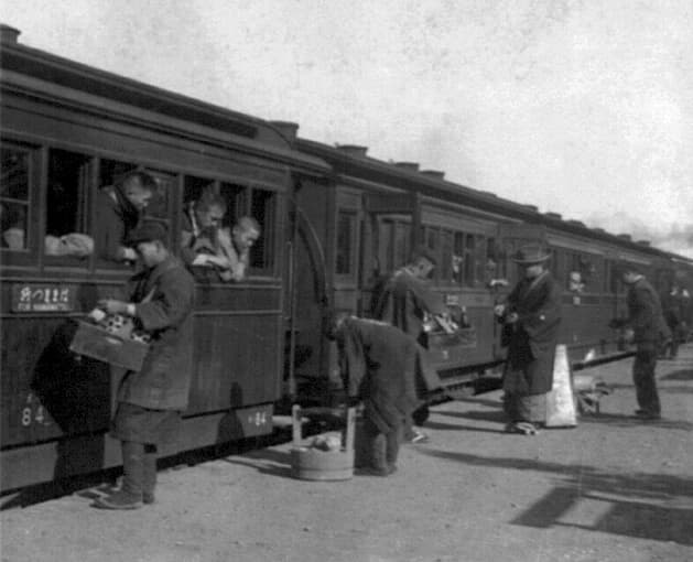 Passengers of train leaning out windows, being served refreshments by men on railroad platform.